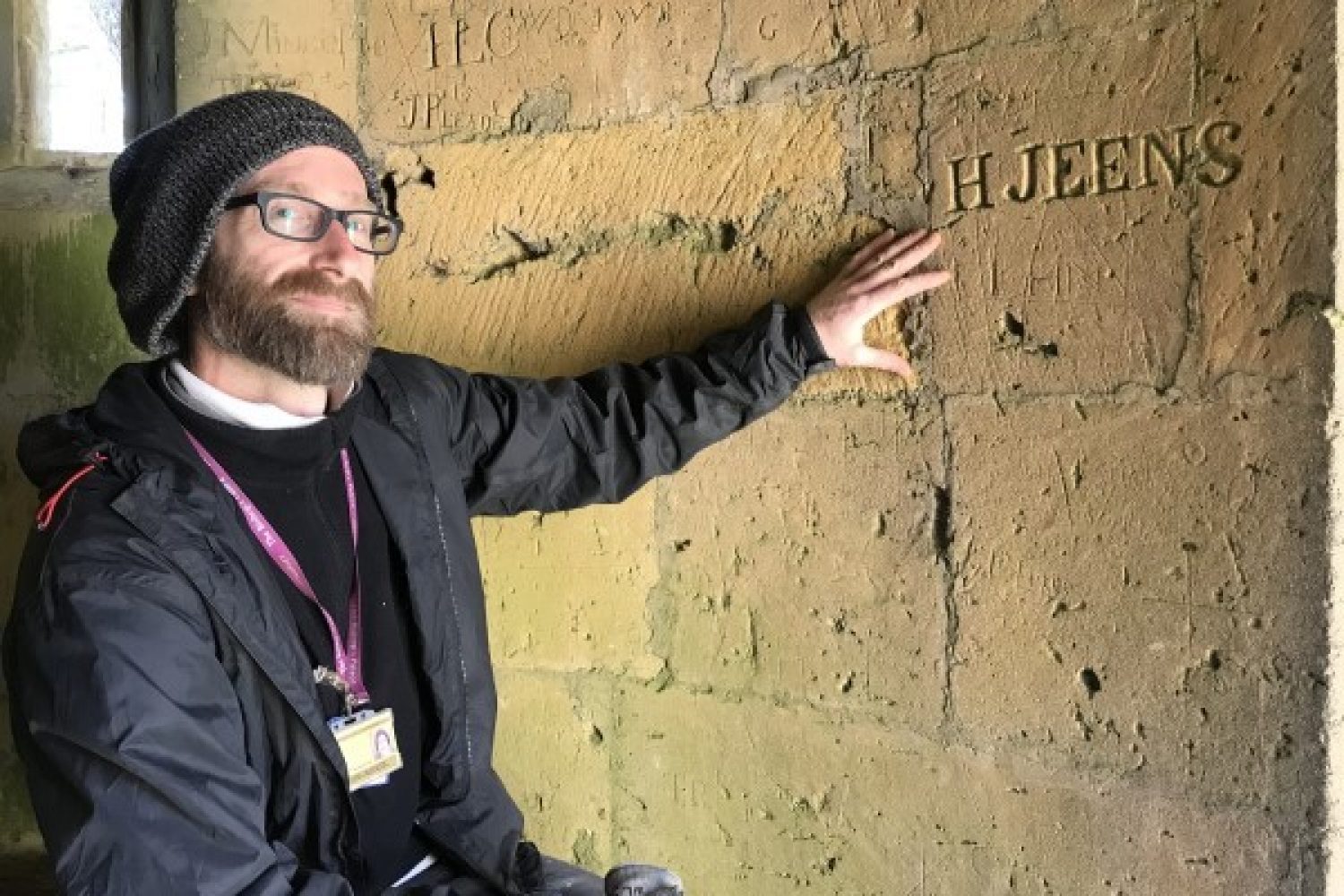 Man standing next to wall that has the text 'H Jeens' deeply carved into it