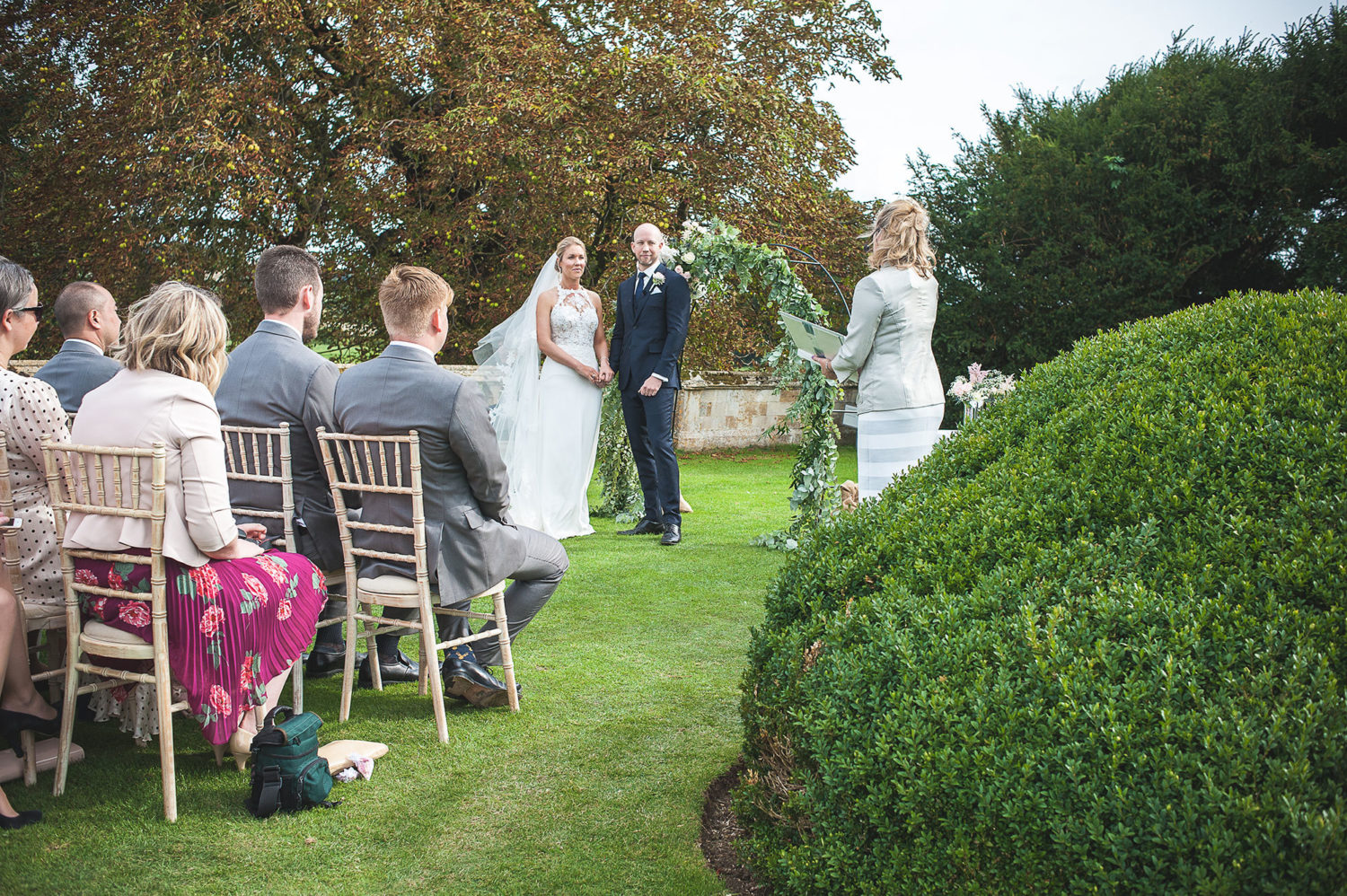 A couple stand hand-in-hand on a lawn in front of seated guests
