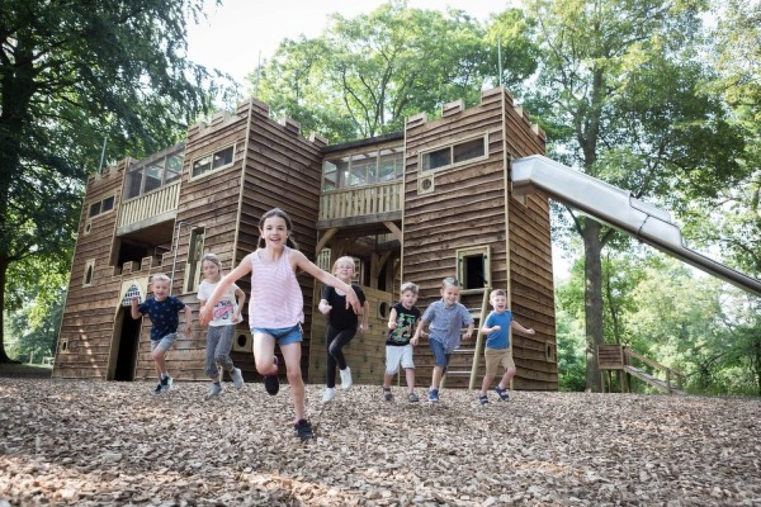 Children running towards the camera in a playground with a fort with a slide in the background