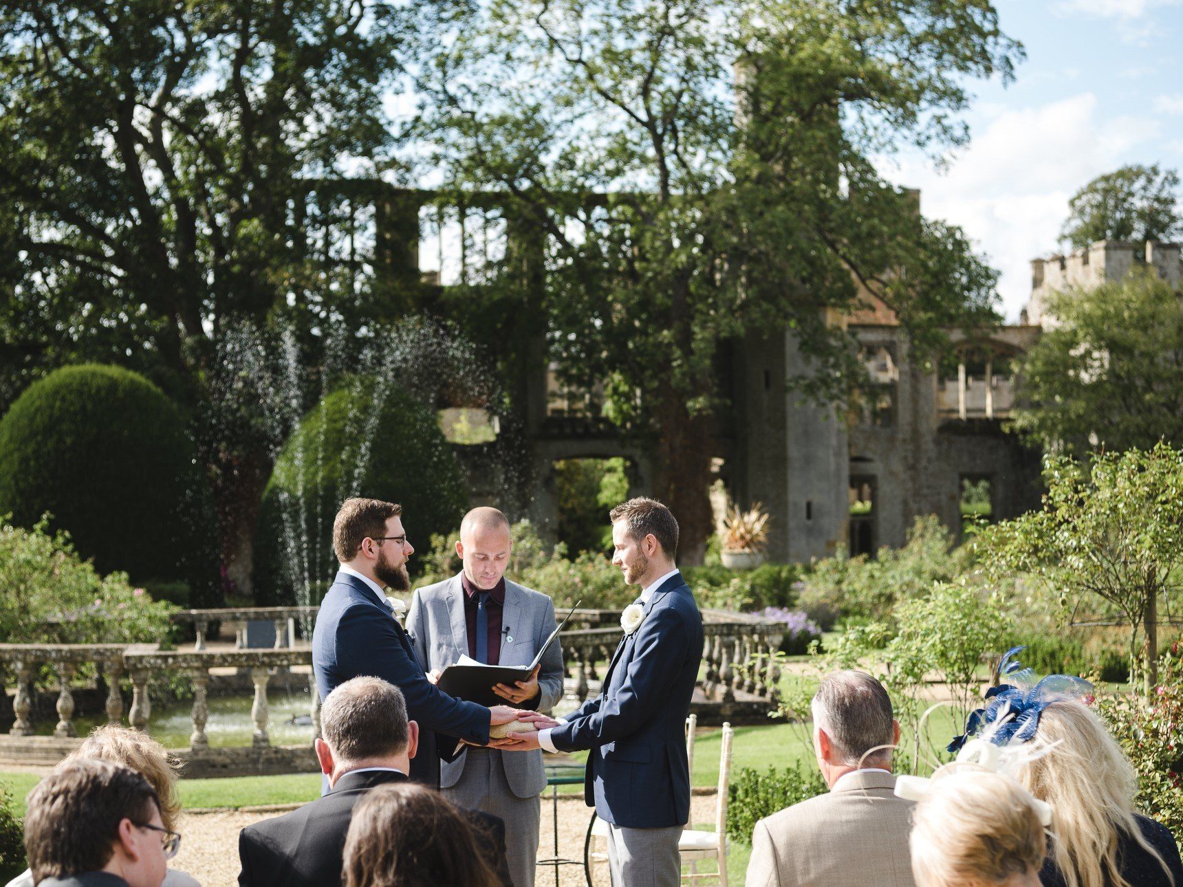 Groomsmen holding hands through wedding vows in the Queen's Garden, with views of castle ruins